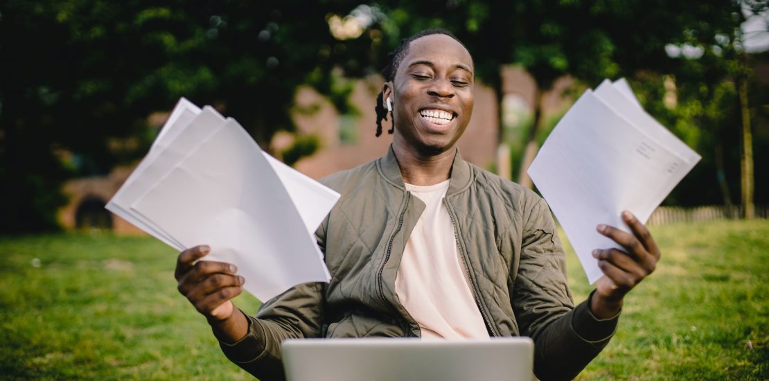 celebrating-success-smiling-man-sat-in-front-of-laptop-with-papers-in-his-hands