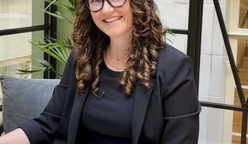 Image of our founder Ellice wearing light beige trousers, black t shirt and black blaxer. she is a white woman crossing her legs, smiling with long curly brown hair and cat eye glasses with black rims.