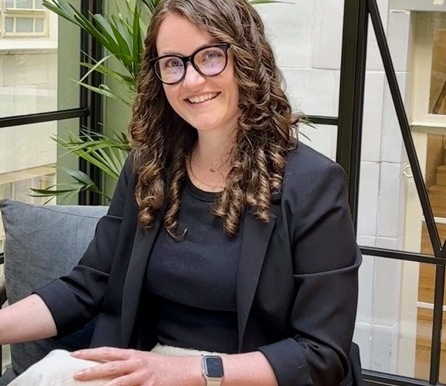 Image of our founder Ellice wearing light beige trousers, black t shirt and black blaxer. she is a white woman crossing her legs, smiling with long curly brown hair and cat eye glasses with black rims.