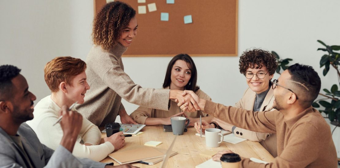 a diverse team of people sat around a table with laptops and coffee. They are all smiling and two are shaking hands.