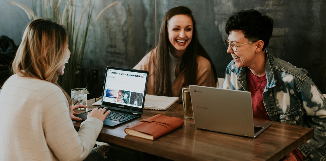 three people sitting around the table with laptops working together, smiling.