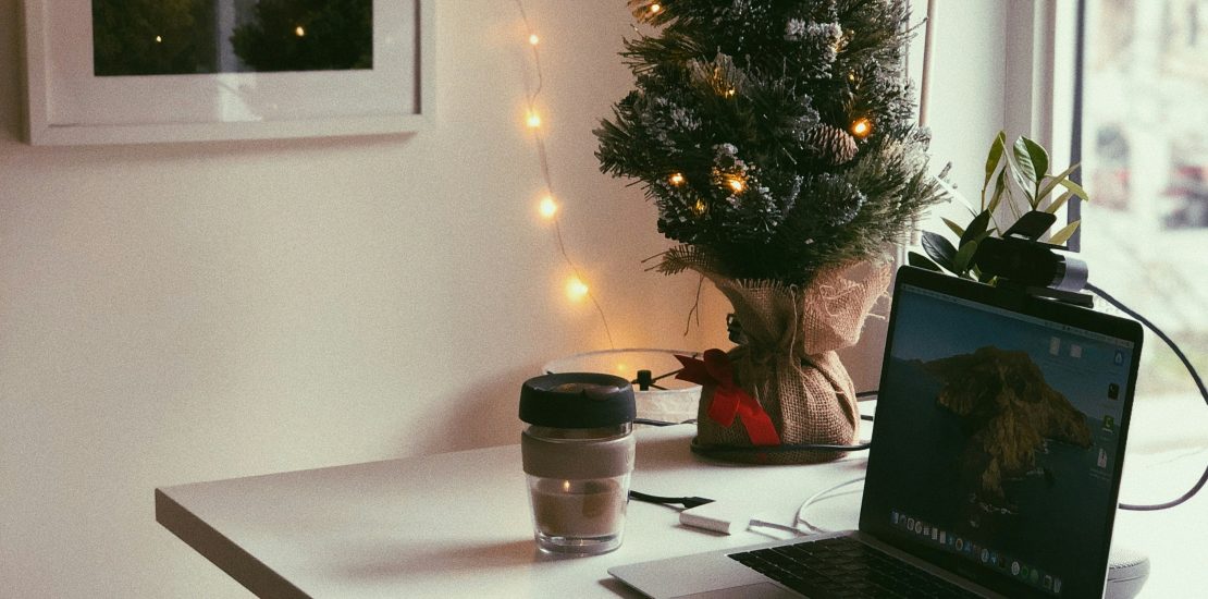 an image of a home workspace with laptop, headphones, notebook and coffee. there is a small christmas tree on the table with fairy lights