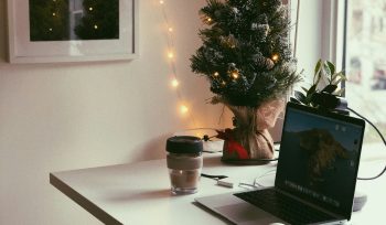 an image of a home workspace with laptop, headphones, notebook and coffee. there is a small christmas tree on the table with fairy lights