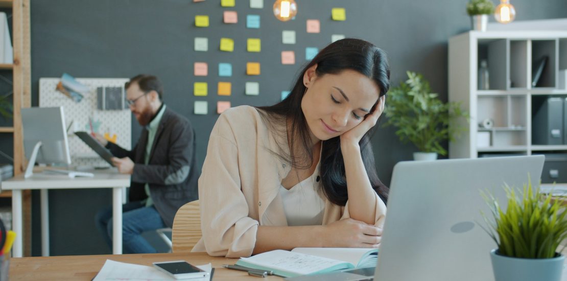 white woman sat at a desk full of paperwork and laptop open. She is leaning on her hand looking tired.