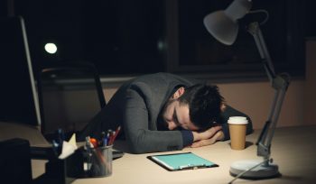 man leaning forward on his desk, asleep with his arms supporting his head. There is a coffee cup and writing pad in front of him.