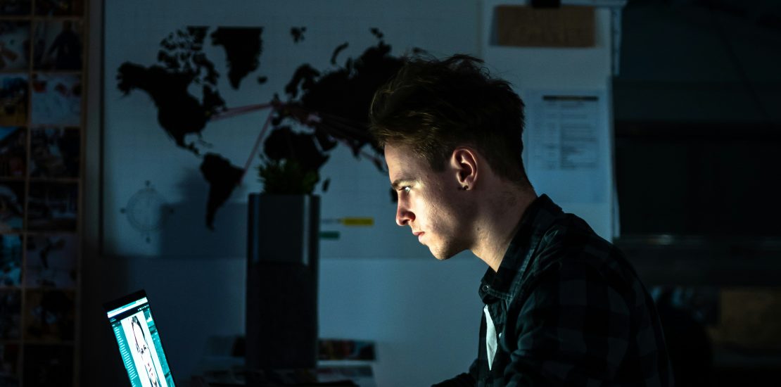 image of a man in a dark room working at his desk on a laptop. the light from the laptop is lighting up his face. Overload