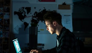 image of a man in a dark room working at his desk on a laptop. the light from the laptop is lighting up his face. Overload