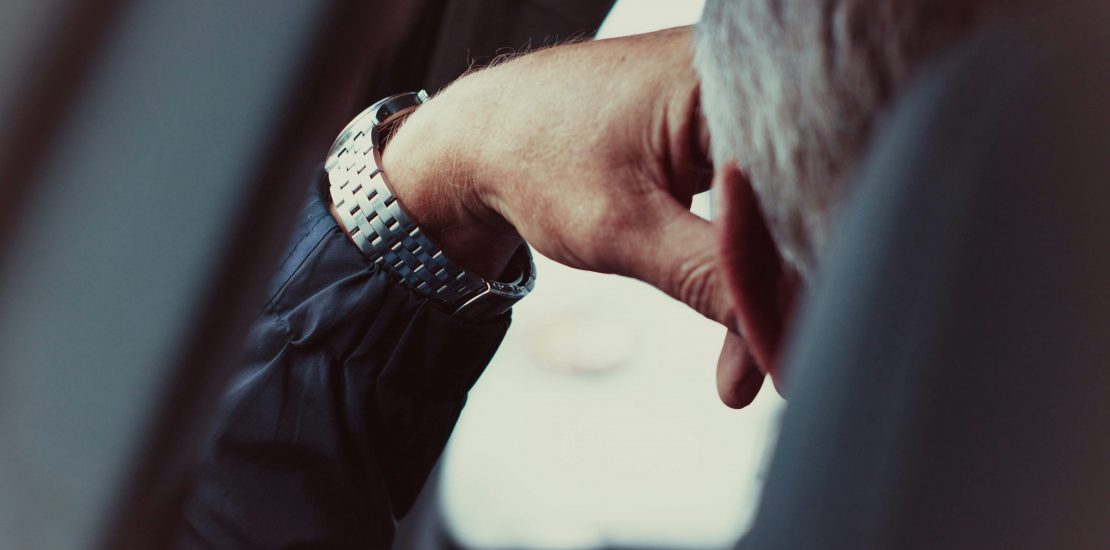 Image is behind the driver of a car with his head resting on his hand. His hair is grey and looks like he is waiting in traffic.