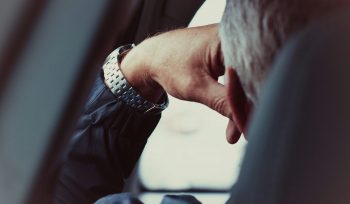 Image is behind the driver of a car with his head resting on his hand. His hair is grey and looks like he is waiting in traffic.
